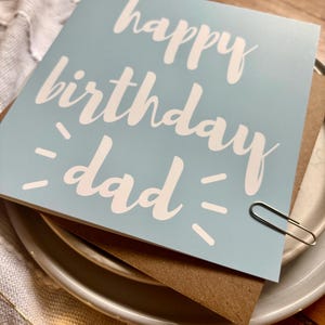 May include: A square light blue greeting card with the white text "happy birthday dad" printed in a script font. The card features small white dash accents on either side of the word "dad" and is secured with a silver metal paper clip.