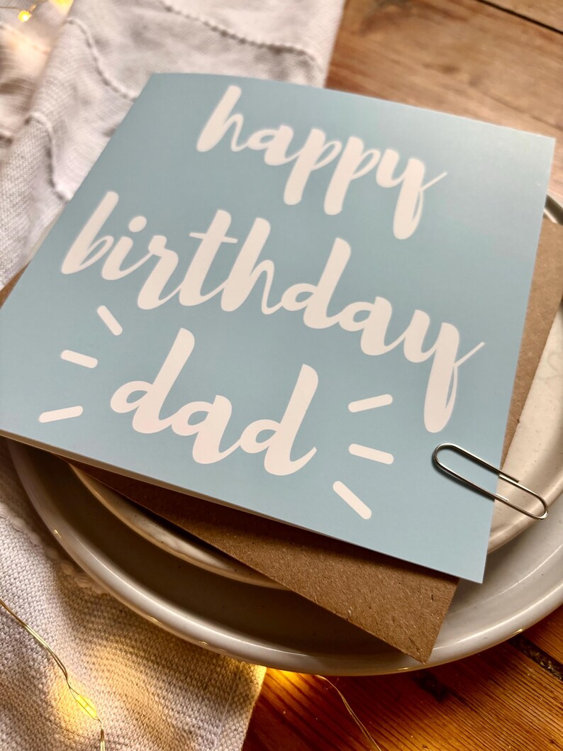 May include: A square light blue greeting card with the white text "happy birthday dad" printed in a script font. The card features small white dash accents on either side of the word "dad" and is secured with a silver metal paper clip.