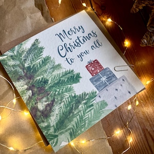 Puede incluir: Una tarjeta de felicitaci&oacute;n cuadrada con una ilustraci&oacute;n en acuarela de un pino verde y una pila de tres regalos envueltos en rojo, azul y blanco. El texto Merry Christmas to you all est&aacute; impreso en letra cursiva azul oscuro sobre el fondo.