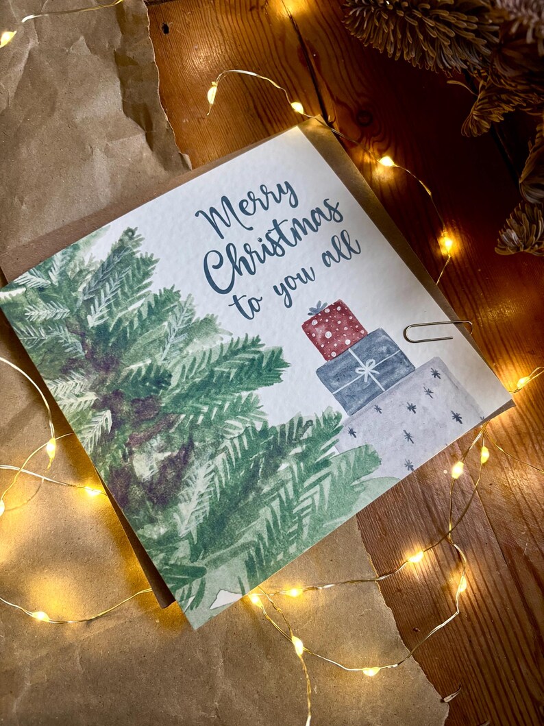 Puede incluir: Una tarjeta de felicitaci&oacute;n cuadrada con una ilustraci&oacute;n en acuarela de un pino verde y una pila de tres regalos envueltos en rojo, azul y blanco. El texto Merry Christmas to you all est&aacute; impreso en letra cursiva azul oscuro sobre el fondo.
