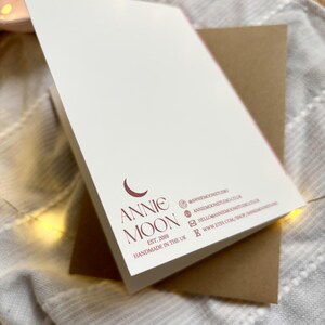 May include: A white card featuring the Annie Moon logo and contact details in burgundy, set against a brown envelope. The card is angled, and the text "Handmade in the UK" is printed below the logo.