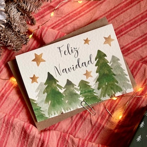 Puede incluir: Una tarjeta de felicitación rectangular con fondo blanco que presenta una ilustración en acuarela de pinos verdes y estrellas doradas. El texto Feliz Navidad está escrito en letra cursiva negra en el centro. La tarjeta está sobre un sobre marrón.