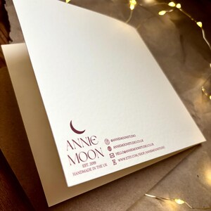 Puede incluir: Una tarjeta de felicitaci&oacute;n plegada de color blanco roto con un logotipo granate oscuro que muestra una luna creciente y el texto ANNIE MOON EST. 2019 HANDMADE IN THE UK. Los datos de contacto incluyen redes sociales, web y correo en granate.