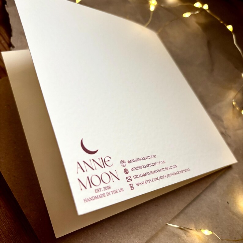 Puede incluir: Una tarjeta de felicitaci&oacute;n plegada de color blanco roto con un logotipo granate oscuro que muestra una luna creciente y el texto ANNIE MOON EST. 2019 HANDMADE IN THE UK. Los datos de contacto incluyen redes sociales, web y correo en granate.