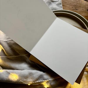 May include: An open greeting card with a blank white page and a light gray page with faint text. The card is on top of a stack of brown cards. The background includes a gray cloth, a tan plate, and a wooden surface with string lights.