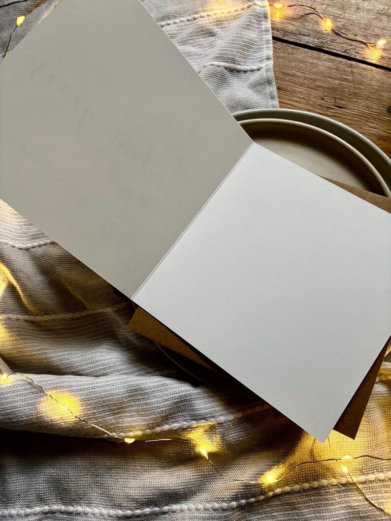 May include: An open greeting card with a blank white page and a light gray page with faint text. The card is on top of a stack of brown cards. The background includes a gray cloth, a tan plate, and a wooden surface with string lights.