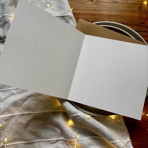 May include: A blank white greeting card opened to reveal a smooth interior surface, accompanied by a brown kraft paper envelope tucked behind it, presented as a clean stationery item for personal messages or custom written notes.
