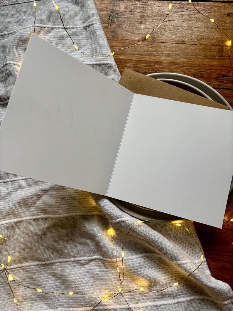 May include: A blank white greeting card opened to reveal a smooth interior surface, accompanied by a brown kraft paper envelope tucked behind it, presented as a clean stationery item for personal messages or custom written notes.