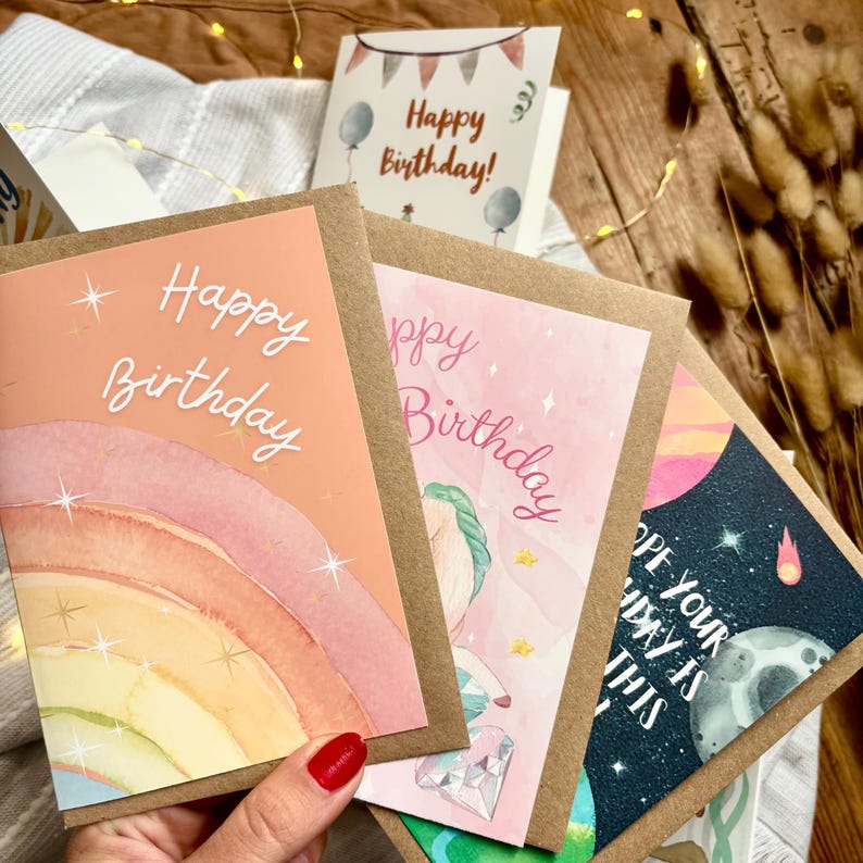 May include: A selection of birthday cards with watercolour illustrations. The cards feature designs with rainbows, unicorns, planets, and the text "Happy Birthday". Each card is in a kraft paper envelope.