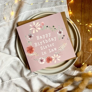 Puede incluir: Una tarjeta de felicitación cuadrada con fondo rosa empolvado que presenta una corona floral de acuarela y el texto blanco "Happy Birthday Sister in Law" centrado. La tarjeta está colocada sobre un sobre marrón y platos blancos.