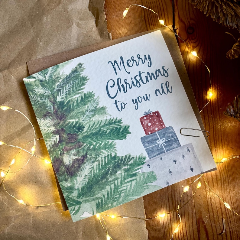 Puede incluir: Una tarjeta de felicitaci&oacute;n cuadrada con una ilustraci&oacute;n en acuarela de un pino verde y una pila de tres regalos envueltos. El texto Merry Christmas to you all est&aacute; impreso en letra cursiva azul oscuro sobre papel marr&oacute;n.