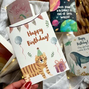 May include: A selection of birthday cards with watercolour illustrations. One card shows a tiger wearing a party hat, balloons, and the text "Happy Birthday!". Other cards feature planets, a whale, and a unicorn.