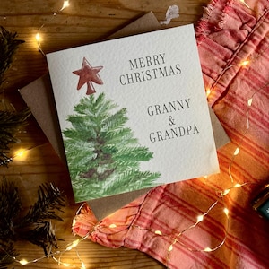 May include: A square greeting card with a watercolor illustration of a green pine tree topped with a red star. The text reads MERRY CHRISTMAS GRANNY & GRANDPA in black lettering. The card is placed on a brown envelope against a festive background.