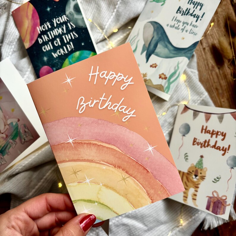 May include: A selection of birthday cards with diverse designs. One card showcases a watercolour rainbow and the words "Happy Birthday." Other cards feature a whale, a tiger, and space-themed graphics. The cards are displayed on a neutral-coloured surface.