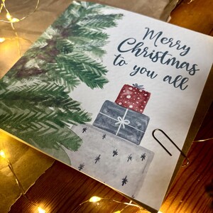 Puede incluir: Una tarjeta de felicitaci&oacute;n cuadrada con una ilustraci&oacute;n en acuarela de un pino verde y una pila de tres regalos envueltos en rojo, azul oscuro y gris claro. El texto Merry Christmas to you all est&aacute; impreso en letra cursiva azul.
