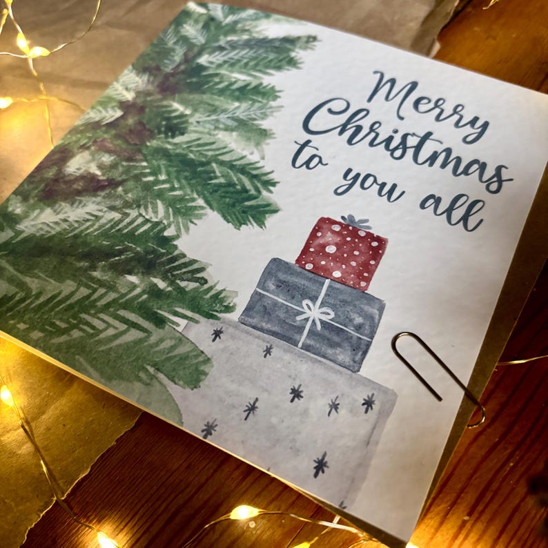Puede incluir: Una tarjeta de felicitaci&oacute;n cuadrada con una ilustraci&oacute;n en acuarela de un pino verde y una pila de tres regalos envueltos en rojo, azul oscuro y gris claro. El texto Merry Christmas to you all est&aacute; impreso en letra cursiva azul.