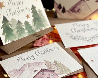Può includere: Una collezione di quattro biglietti d'auguri bianchi in carta ruvida, ognuno con illustrazioni ad acquerello di scene invernali, tra cui pini, una baita di montagna e uno skilift, tutti con il testo scritto a mano Merry Christmas.