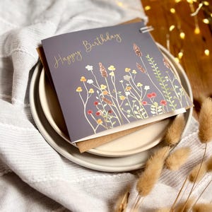 May include: A gray birthday card with gold lettering that reads "Happy Birthday." The card features a watercolor illustration of wildflowers in shades of yellow, orange, and red. The card is on a white plate with a brown envelope.