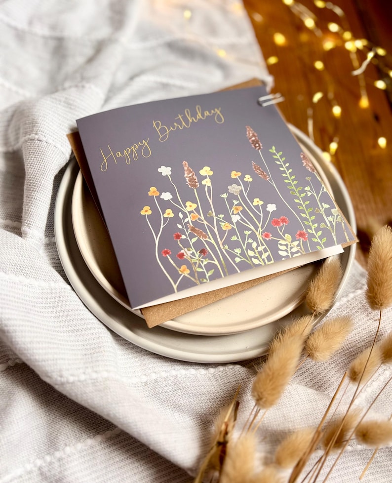 May include: A gray birthday card with gold lettering that reads "Happy Birthday." The card features a watercolor illustration of wildflowers in shades of yellow, orange, and red. The card is on a white plate with a brown envelope.