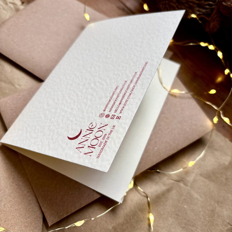 Pu&ograve; includere: Un biglietto d'auguri piegato bianco con una superficie testurizzata che presenta un testo rosso scuro che recita ANNIE MOON, EST. 2019, HANDMADE IN THE UK, insieme a profili social, un indirizzo email e un URL del sito web sul retro.