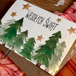 Pu&ograve; includere: Un biglietto d'auguri rettangolare con un'illustrazione ad acquerello di alberi di pino verdi e stelle dorate su sfondo bianco. Il testo WESOŁYCH ŚWIĄT &egrave; stampato in inchiostro nero al centro. Una graffetta metallica &egrave; fissata sul lato.