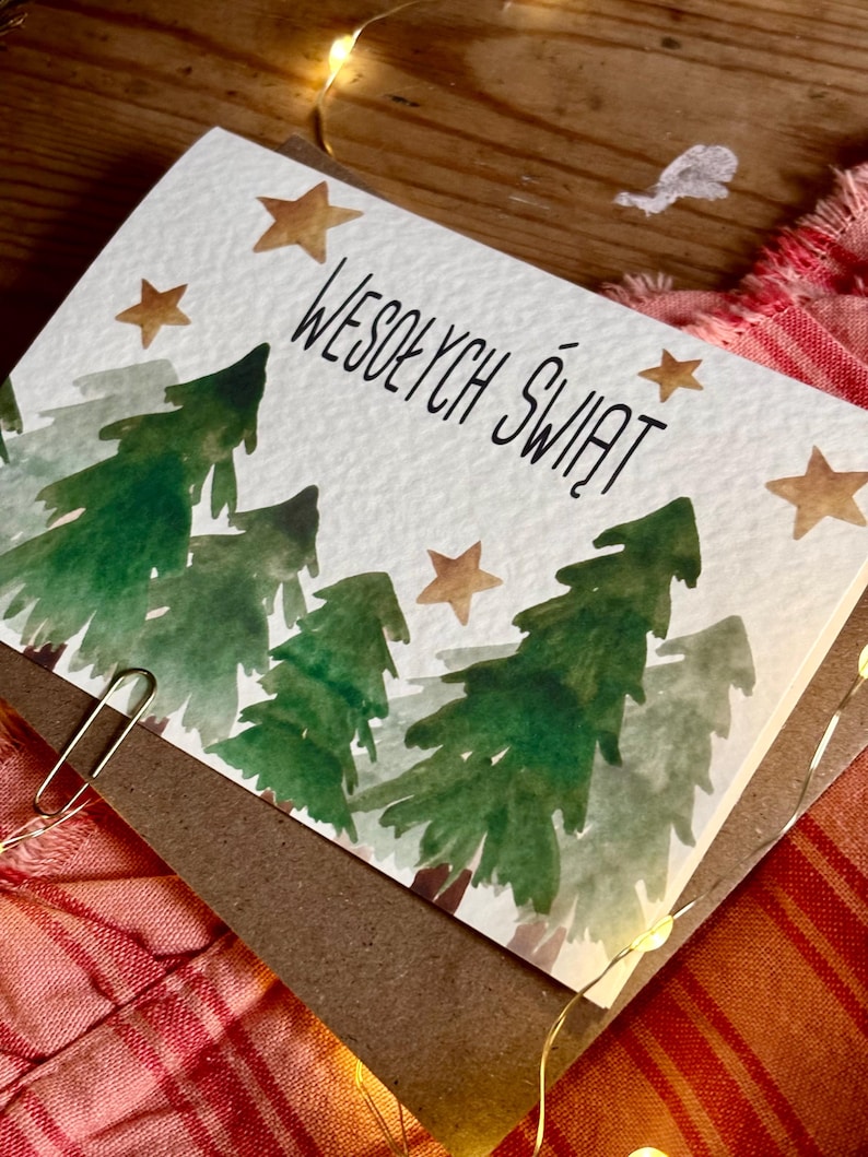 Pu&ograve; includere: Un biglietto d'auguri rettangolare con un'illustrazione ad acquerello di alberi di pino verdi e stelle dorate su sfondo bianco. Il testo WESOŁYCH ŚWIĄT &egrave; stampato in inchiostro nero al centro. Una graffetta metallica &egrave; fissata sul lato.