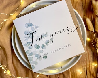 May include: A square greeting card with a white background featuring a watercolor illustration of green eucalyptus leaves. The text reads "Ten years" in a black script font and "HAPPY ANNIVERSARY" in a simple black sans-serif typeface.