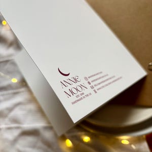 May include: A white greeting card with the text "Annie Moon" in burgundy script. The card is open, revealing a blank interior. The card is handmade in the UK.