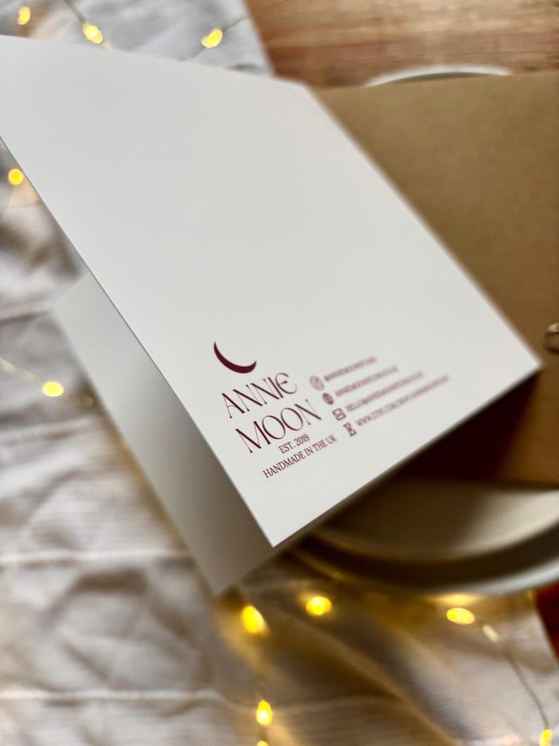 May include: A white greeting card with the text "Annie Moon" in burgundy script. The card is open, revealing a blank interior. The card is handmade in the UK.