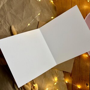 Puede incluir: Una tarjeta de felicitaci&oacute;n blanca en blanco abierta para revelar dos p&aacute;ginas interiores vac&iacute;as. La tarjeta est&aacute; colocada en &aacute;ngulo, mostrando su superficie blanca mate limpia y una l&iacute;nea de pliegue n&iacute;tida en el centro, ofreciendo un espacio liso.