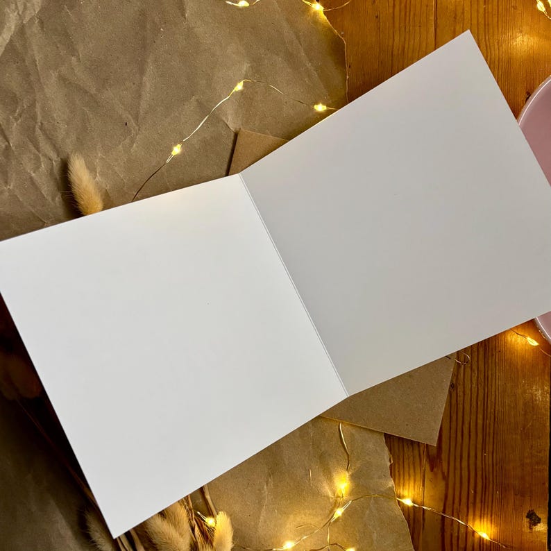 Puede incluir: Una tarjeta de felicitaci&oacute;n blanca en blanco abierta para revelar dos p&aacute;ginas interiores vac&iacute;as. La tarjeta est&aacute; colocada en &aacute;ngulo, mostrando su superficie blanca mate limpia y una l&iacute;nea de pliegue n&iacute;tida en el centro, ofreciendo un espacio liso.