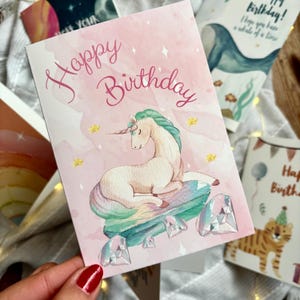 May include: A pink birthday card featuring a unicorn illustration. The card says "Happy Birthday" in cursive. The unicorn rests on a gem. Other cards with illustrations of a whale and a tiger are visible in the background.