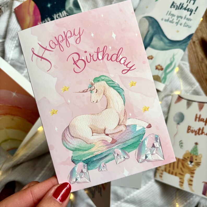 May include: A pink birthday card featuring a unicorn illustration. The card says "Happy Birthday" in cursive. The unicorn rests on a gem. Other cards with illustrations of a whale and a tiger are visible in the background.