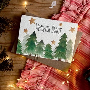 Pu&ograve; includere: Un biglietto d'auguri rettangolare con un'illustrazione ad acquerello di alberi di pino verdi e stelle dorate. Il testo WESOŁYCH ŚWIĄT &egrave; stampato in nero al centro. Una graffetta argentata &egrave; fissata sul bordo inferiore del biglietto.