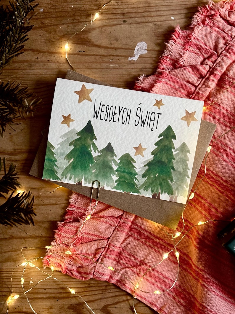 Pu&ograve; includere: Un biglietto d'auguri rettangolare con un'illustrazione ad acquerello di alberi di pino verdi e stelle dorate. Il testo WESOŁYCH ŚWIĄT &egrave; stampato in nero al centro. Una graffetta argentata &egrave; fissata sul bordo inferiore del biglietto.