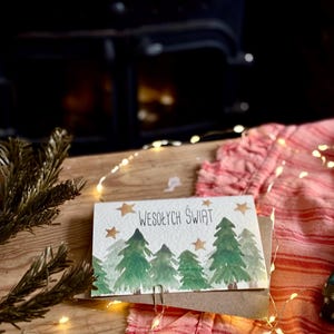 Pu&ograve; includere: Un biglietto d'auguri rettangolare con un'illustrazione ad acquerello di pini verdi e stelle dorate su sfondo bianco. Il testo WESOŁYCH ŚWIĄT &egrave; stampato in nero al centro. Il biglietto &egrave; fissato con una graffetta metallica a una busta marrone.