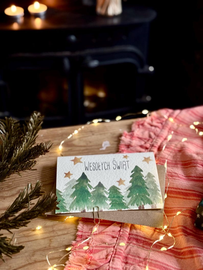 Pu&ograve; includere: Un biglietto d'auguri rettangolare con un'illustrazione ad acquerello di pini verdi e stelle dorate su sfondo bianco. Il testo WESOŁYCH ŚWIĄT &egrave; stampato in nero al centro. Il biglietto &egrave; fissato con una graffetta metallica a una busta marrone.