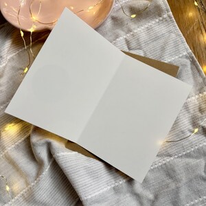 May include: An open, blank greeting card with a white interior, resting on a grey and white striped fabric. The card is paired with a brown envelope. A pink plate and string lights are in the background, suggesting a festive or celebratory theme.
