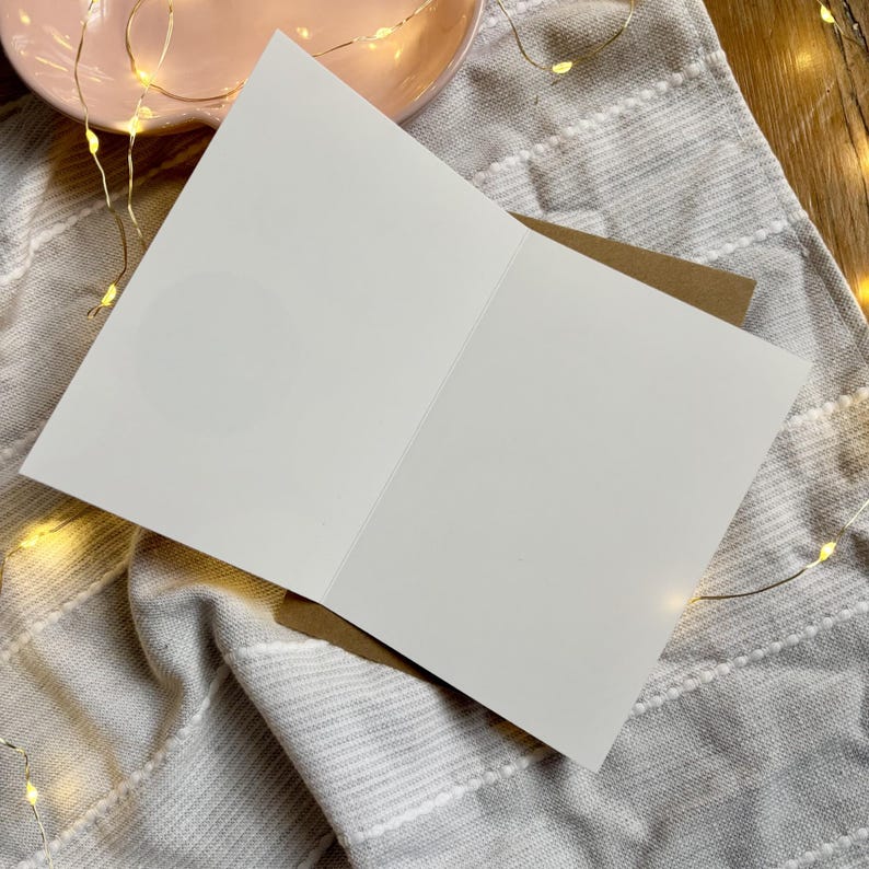 May include: An open, blank greeting card with a white interior, resting on a grey and white striped fabric. The card is paired with a brown envelope. A pink plate and string lights are in the background, suggesting a festive or celebratory theme.