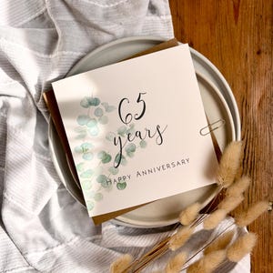 May include: A square greeting card with a white background featuring a watercolor illustration of green eucalyptus leaves on the left side. The text reads 65 years in black script and HAPPY ANNIVERSARY in black capital letters below it.