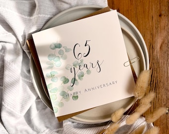 May include: A square greeting card with a white background featuring a watercolor illustration of green eucalyptus leaves on the left side. The text reads 65 years in black script and HAPPY ANNIVERSARY in black capital letters below it.