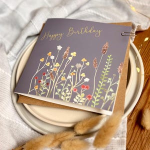 May include: A gray birthday card with the words "Happy Birthday" in gold script. The card features a watercolor illustration of wildflowers in shades of red, yellow, white, and green. The card is held together with a silver paper clip.