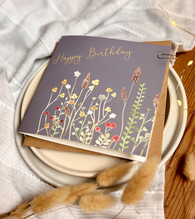 May include: A gray birthday card with the words "Happy Birthday" in gold script. The card features a watercolor illustration of wildflowers in shades of red, yellow, white, and green. The card is held together with a silver paper clip.