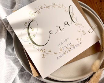 May include: A white card with the word "Coral" in elegant script, surrounded by a floral wreath. The card reads "Happy 35th Anniversary." It rests on a plate with a brown envelope and a paper clip.