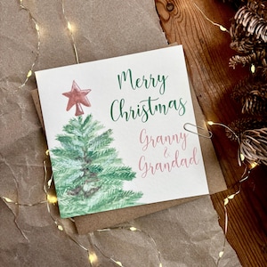 May include: A Christmas card with a watercolor illustration of a green Christmas tree topped with a red star. The card reads "Merry Christmas Granny & Grandad" in green and pink script.