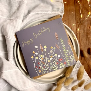 May include: A gray birthday card with gold lettering and a floral design. The card is clipped to a brown backing and rests on a white plate atop a gray cloth. Dried flowers are in the foreground.