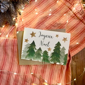 Puede incluir: Una tarjeta de felicitación rectangular blanca con una ilustración en acuarela de pinos verdes y estrellas doradas con el texto Joyeux Noel escrito en letra cursiva negra. La tarjeta está colocada sobre un sobre marrón.