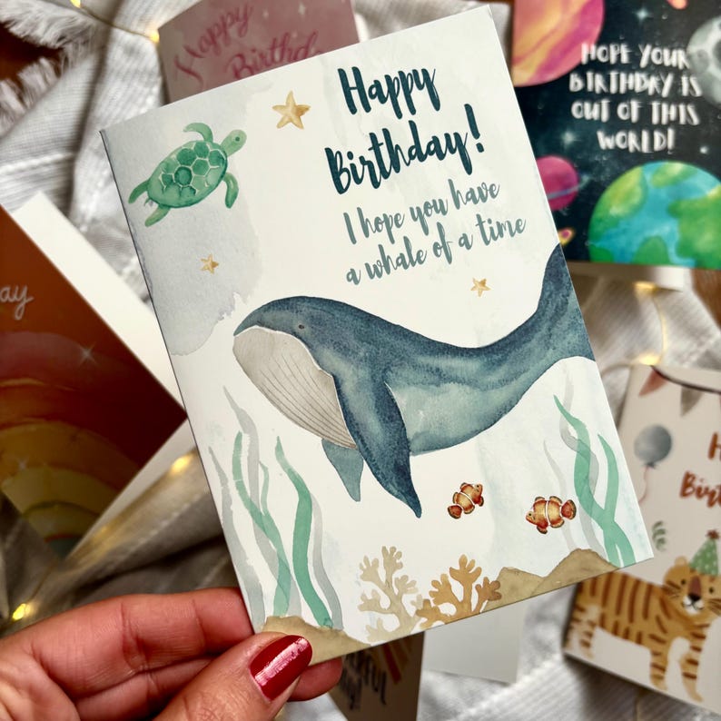 May include: A birthday card featuring a watercolour illustration of a blue whale, a green turtle, and two clownfish. The card reads "Happy Birthday! I hope you have a whale of a time." Other cards are visible in the background.