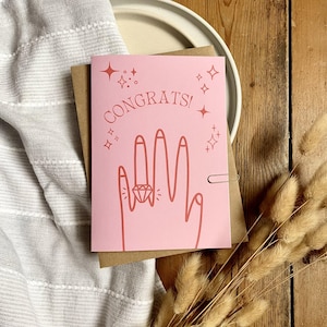 Tarjeta de compromiso en rosa y rojo con mano y anillo. Romántica y moderna tarjeta de felicitación para recién comprometidos, E6