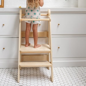 May include: A child stands on a wooden step stool in a kitchen. The stool has two steps and a safety rail. The child is wearing a white and green dress and has long brown hair.
